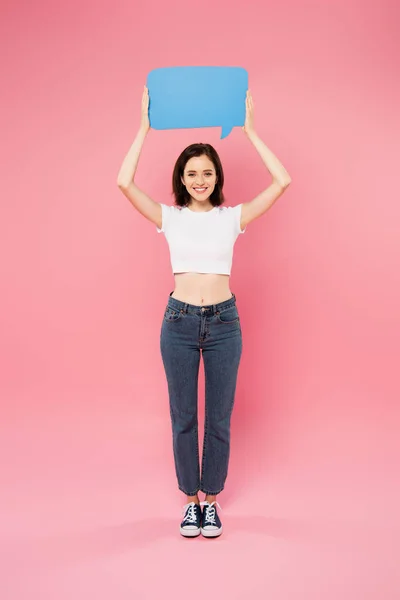 Vista de longitud completa de sonriente chica bonita sosteniendo en blanco burbuja de habla azul aislado en rosa - foto de stock