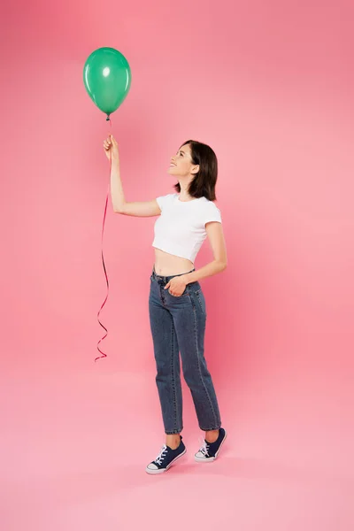 Full length view of smiling pretty girl looking at green balloon isolated on pink — Stock Photo