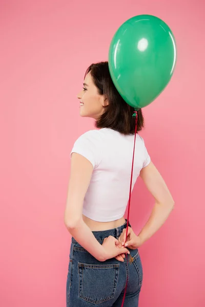Sorridente bella ragazza tenendo palloncino verde dietro la schiena isolato su rosa — Foto stock