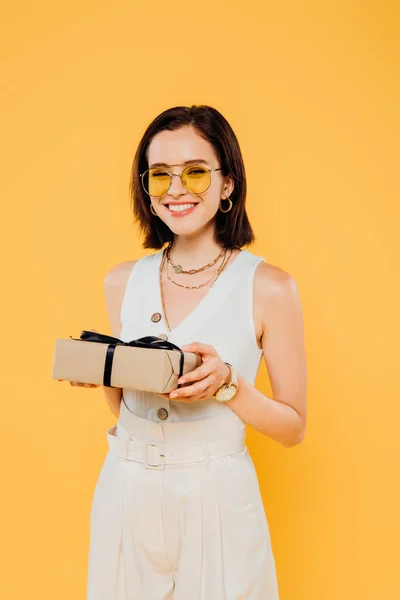 Улыбающаяся элегантная женщина в солнечных очках с подарочной коробкой, изолированной на желтый — стоковое фото