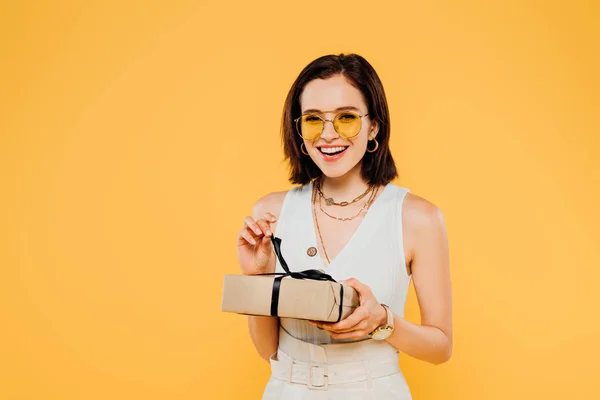 Aufgeregt lächelnde elegante Frau mit Sonnenbrille hält Geschenkbox isoliert auf gelb — Stockfoto