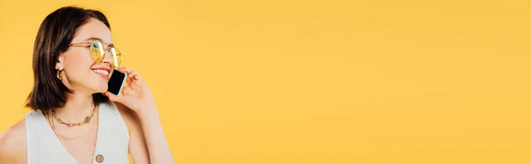 Plan panoramique de souriante femme élégante en lunettes de soleil parlant sur smartphone isolé sur jaune — Photo de stock
