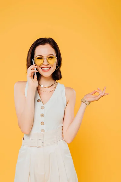 Усміхнена елегантна жінка в сонцезахисних окулярах говорить на смартфоні ізольовано на жовтому — стокове фото