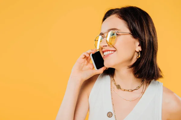 Усміхнена елегантна жінка в сонцезахисних окулярах говорить на смартфоні ізольовано на жовтому — стокове фото