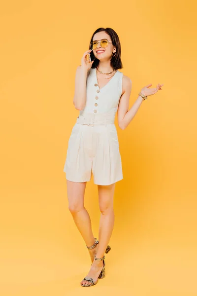 Full length view of smiling elegant woman in sunglasses talking on smartphone isolated on yellow — Stock Photo