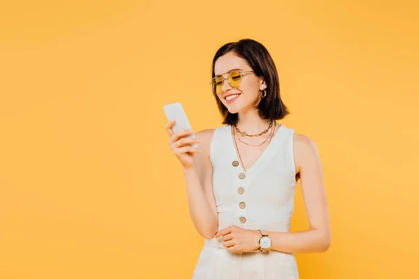 Mujer elegante sonriente en gafas de sol usando teléfono inteligente aislado en amarillo - foto de stock