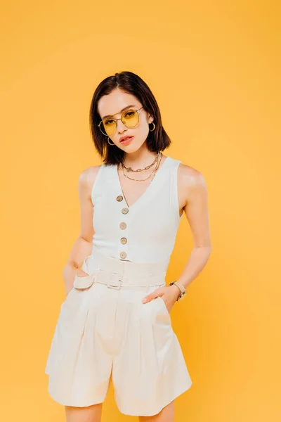 Femme élégante dans des lunettes de soleil posant avec les mains dans des poches isolées sur jaune — Photo de stock