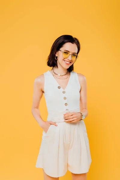 Smiling stylish woman in sunglasses posing with hand in pocket isolated on yellow — Stock Photo
