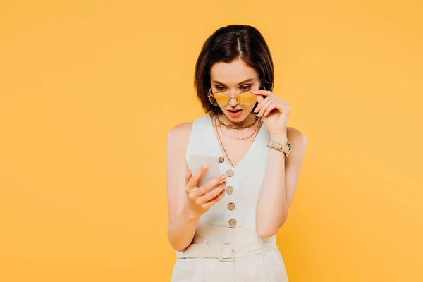 Femme choquée en lunettes de soleil regardant smartphone et lunettes de soleil touchantes isolées sur jaune — Photo de stock