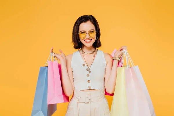 Femme souriante dans des lunettes de soleil tenant des sacs à provisions isolés sur jaune — Photo de stock