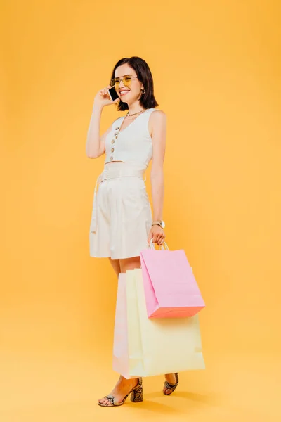 Piena lunghezza vista di donna felice sorridente in occhiali da sole tenendo borse della spesa e parlando su smartphone isolato su giallo — Foto stock