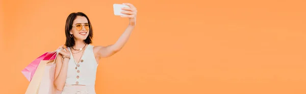 Menina na moda feliz em óculos de sol com sacos de compras tomando selfie no smartphone isolado em laranja — Fotografia de Stock