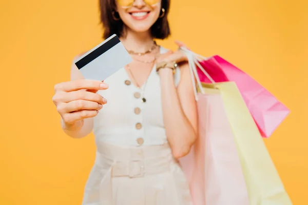 Vista recortada de chica feliz con bolsas de compras presentando tarjeta de crédito aislada en amarillo - foto de stock