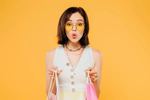 Chocado chica de moda en gafas de sol sosteniendo bolsas de compras aisladas en amarillo - foto de stock