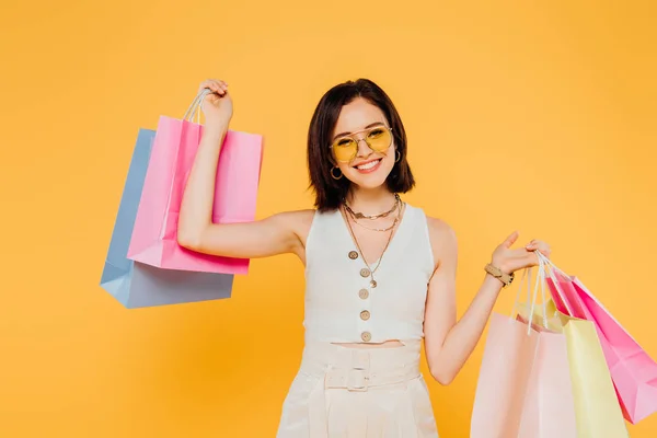 Усміхнена модна дівчина в сонцезахисних окулярах тримає сумки для покупок ізольовані на жовтому — стокове фото