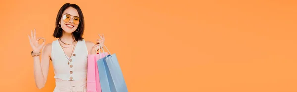 Счастливая модная девушка в солнечных очках, держа сумки и показывая хорошо знак изолирован на оранжевый — стоковое фото