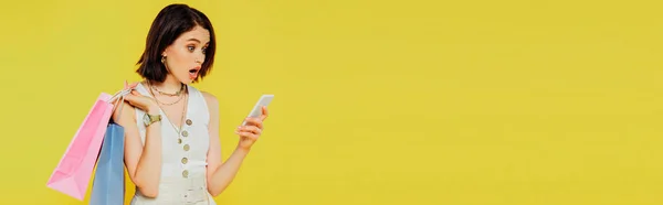 Tiro panorâmico de menina chocada com sacos de compras falando no smartphone isolado no amarelo — Fotografia de Stock