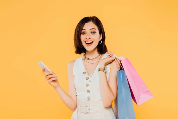 Excitada chica de moda con bolsas de compras hablando en el teléfono inteligente aislado en amarillo - foto de stock