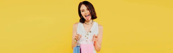 Panoramic shot of fashionable girl with shopping bags showing tongue isolated on yellow — Stock Photo
