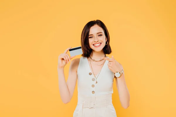 Smiling elegant girl pointing with finger at credit card isolated on yellow — Stock Photo