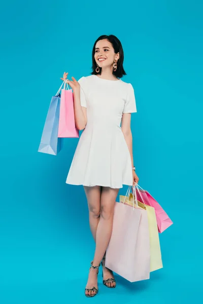 Vista de longitud completa de la mujer elegante sonriente en vestido con bolsas de compras mirando hacia otro lado aislado en azul - foto de stock