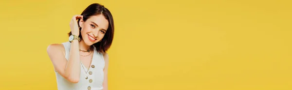 Панорамний знімок усміхненої елегантної дівчини торкається волосся ізольовано на жовтому — стокове фото