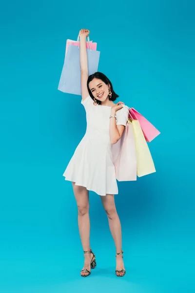 Visão comprimento total de mulher elegante sorridente no vestido com sacos de compras isolados em azul — Fotografia de Stock