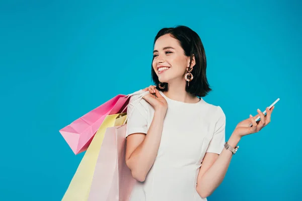 Sorrindo mulher elegante no vestido com sacos de compras e smartphone olhando para longe isolado em azul — Fotografia de Stock