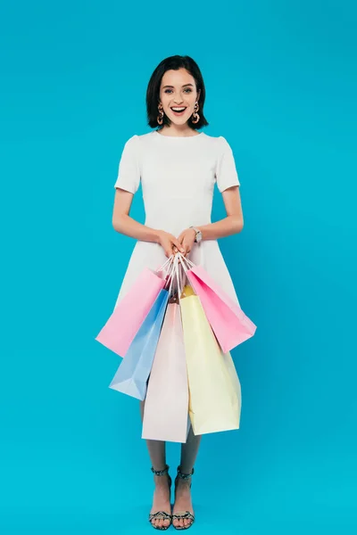 Ganzkörperansicht einer lächelnden eleganten Frau im Kleid mit Einkaufstaschen auf blauem Grund — Stockfoto
