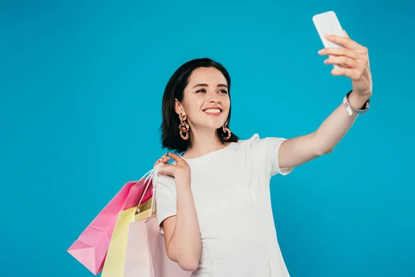 Sonriente mujer elegante en vestido con bolsas de compras tomando selfie en el teléfono inteligente aislado en azul - foto de stock