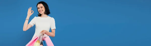 Панорамный снимок улыбающейся элегантной женщины в платье с пакетами для покупок, показывающих знак ОК изолированы на синий — стоковое фото