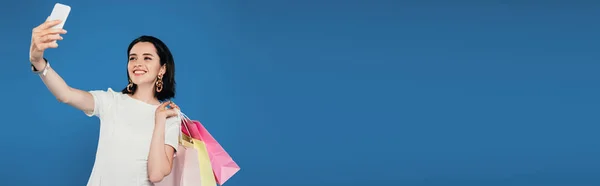 Plano panorámico de mujer elegante sonriente en vestido con bolsas de compras tomando selfie en el teléfono inteligente aislado en azul - foto de stock