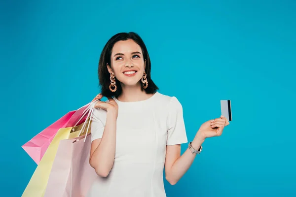 Усміхнена елегантна жінка в одязі з сумками тримає кредитну картку ізольовано на синьому — стокове фото