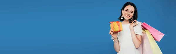 Colpo panoramico di donna elegante sorridente in abito con borse della spesa in possesso di scatola regalo isolato su blu — Foto stock