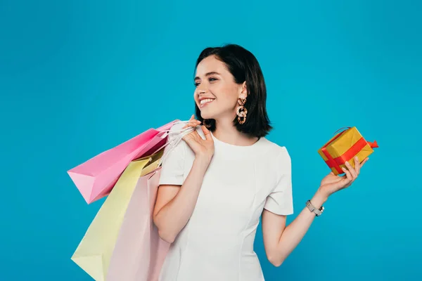 Mujer elegante sonriente en vestido con bolsas de compras sosteniendo caja de regalo y mirando hacia otro lado aislado en azul - foto de stock