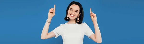 Panoramic shot of smiling elegant girl pointing with fingers upwards isolated on blue — Stock Photo