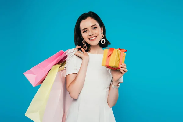 Sonriente mujer elegante en vestido sosteniendo bolsas de compras y caja de regalo aislado en azul - foto de stock