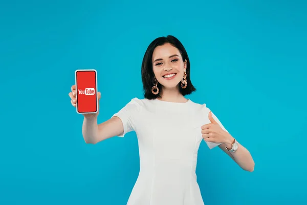 KYIV, UCRAINA - 3 LUGLIO 2019: donna elegante e sorridente in abito che tiene lo smartphone con logo youtube e mostra il pollice isolato sul blu — Foto stock