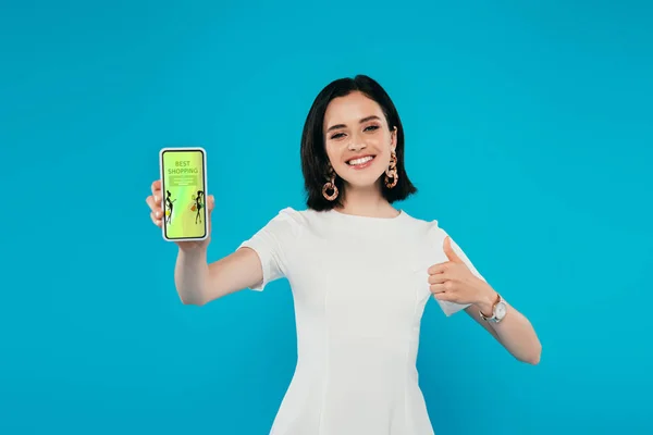 Sonriente mujer elegante en vestido sosteniendo teléfono inteligente con la aplicación de compras de moda y mostrando el pulgar hacia arriba aislado en azul - foto de stock