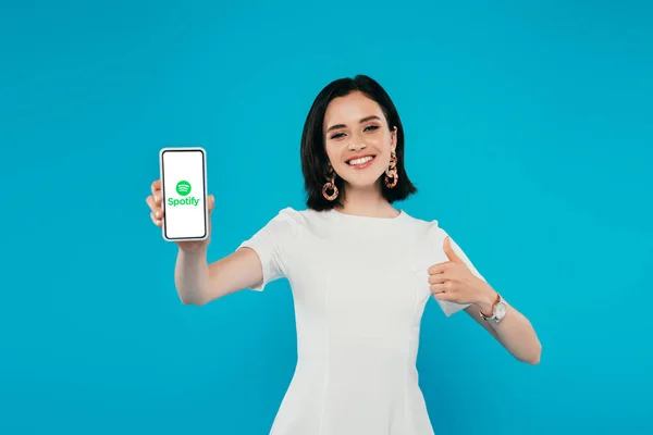 KYIV, UKRAINE - JULY 3, 2019: smiling elegant woman in dress holding smartphone with spotify logo and showing thumb up isolated on blue — Stock Photo