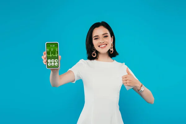 Sonriente mujer elegante en vestido sosteniendo teléfono inteligente con aplicación sanitaria y mostrando el pulgar hacia arriba aislado en azul - foto de stock