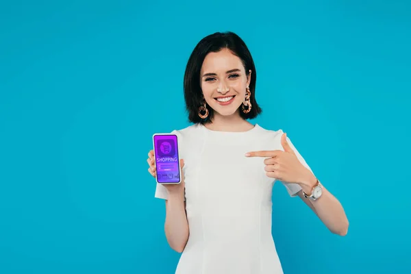 Sonriente mujer elegante en vestido apuntando con el dedo en el teléfono inteligente con aplicación de compras en línea aislado en azul - foto de stock