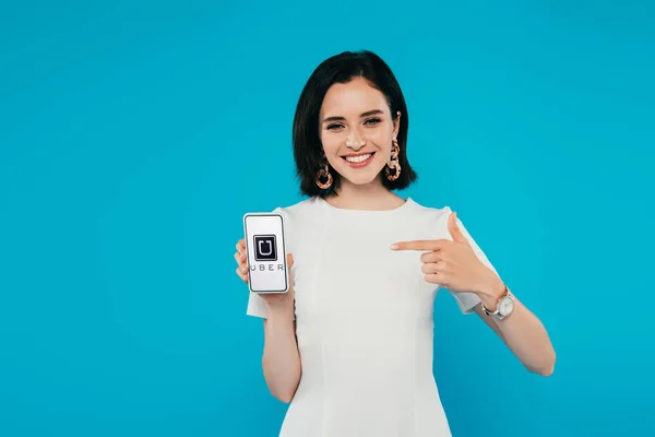 KYIV, UKRAINE - JULHO 3, 2019: sorrindo mulher elegante em vestido apontando com o dedo para o smartphone com logotipo uber isolado em azul — Fotografia de Stock