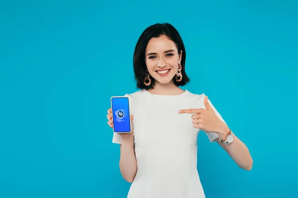KYIV, UCRAINA - 3 LUGLIO 2019: donna elegante e sorridente in abito che punta con il dito lo smartphone con logo shazam isolato sul blu — Foto stock