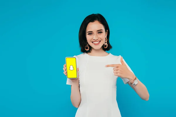 KYIV, UKRAINE - JULHO 3, 2019: sorrindo mulher elegante em vestido apontando com o dedo para o smartphone com logotipo Snapchat isolado em azul — Fotografia de Stock