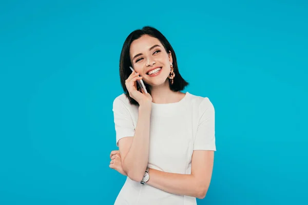 Sonriente mujer elegante en vestido hablando en el teléfono inteligente y mirando hacia otro lado aislado en azul - foto de stock