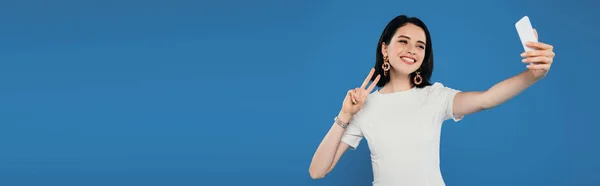 Colpo panoramico di donna elegante sorridente in abito prendendo selfie e mostrando segno di pace isolato su blu — Foto stock