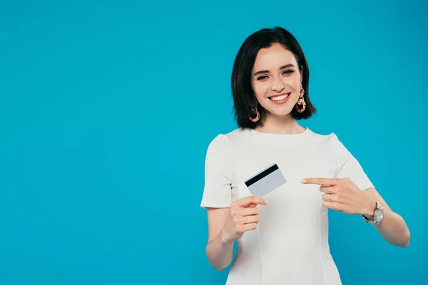 Sonriente mujer elegante en vestido apuntando con el dedo a la tarjeta de crédito aislado en azul - foto de stock