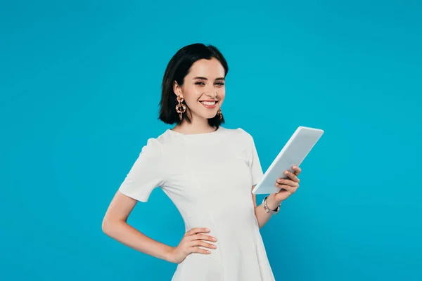 Lächelnde elegante Frau im Kleid mit der Hand auf der Hüfte, die ein digitales Tablet hält, isoliert auf blau — Stockfoto