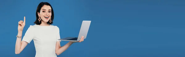 Panoramic shot of excited smiling elegant woman holding laptop and showing idea gesture isolated on blue — Stock Photo